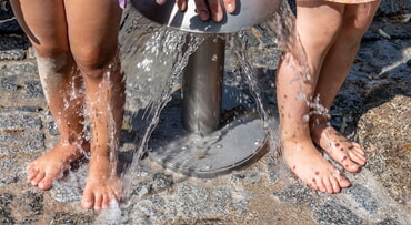 Kinderbeine unter einem Wasserspender im Freien. Wasser flie&szlig;t auf steinernen Boden.