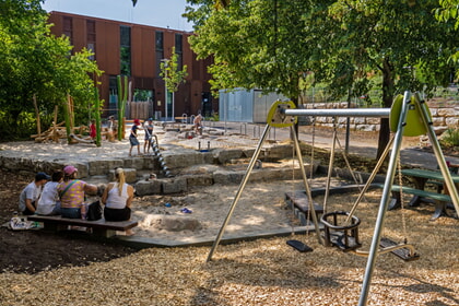 Spielplatz mit Schaukeln, Sandkasten und Sitzgelegenheiten. Erwachsene sitzen und Kinder spielen im Freien.