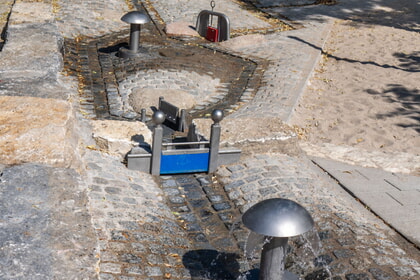 Wasserspielplatz mit mehreren blauen und grauen Wasserspr&uuml;hern auf gepflastertem Untergrund.